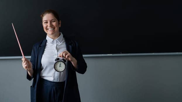 Caucasian female teacher in a pantsuit holds a pointer and a school alarm clock near the blackboard