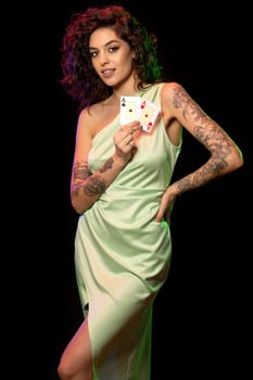 Confident smiling stylish young female poker player standing against dark background, holding pair of aces with winner look. Gambling concept