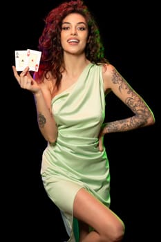 Smiling gambling attractive young woman in silk green dress with tattoo on arms holding winning set of two aces cards posing against black background. Poker game concept