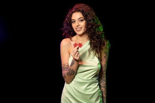 Portrait of smiling young woman in light green dress with tattoo on arms showing two red betting tokens, standing on dark studio background. Casino game concept