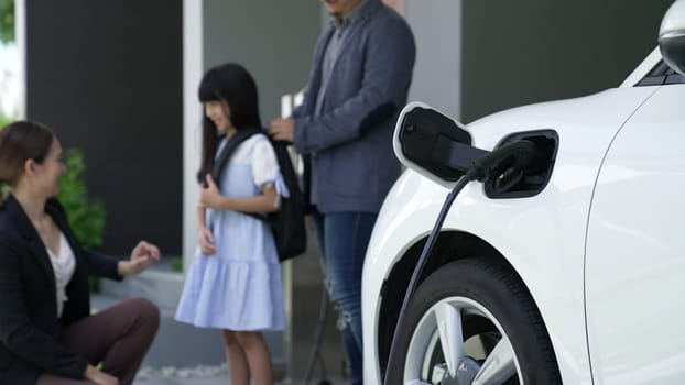 Progressive parents with electric vehicle and home charging station. Happy family with daughter giving each other high fives before leave for school. Alternative future transportation concept of EVs