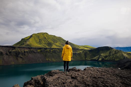 his photo shows a backpacker girl contemplating the beauty of Blahylur Lake in Iceland. She is standing in the center of the frame, wearing a yellow trekking jacket and a grey hat. The sky is overcast, creating a dramatic and atmospheric scene. The lake is a stunning blue-green color, and the green mossy mountain in the background. This photo is perfect for use in travel and nature photography. It could be used to illustrate a story about hiking in Iceland, or it could simply be used to create a sense of contemplation and reflection.
