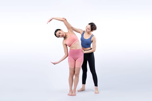 Asian woman in sportswear doing yoga exercise in standing pose with her trainer or yoga buddy. Healthy body care and meditation yoga lifestyle in full shot on isolated background. Vigorous