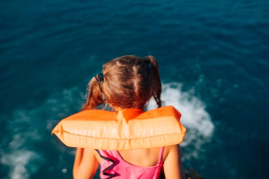 A young girl wearing a bright life jacket is gazing at the serene blue water, feeling ready for an exciting adventure
