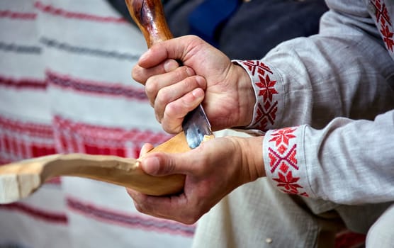 A craftsman, dressed in traditional attire, carefully carves a wooden spoon with a chisel and mallet. He is seated on a wooden stump, surrounded by the remnants of his work and the vibrant colors of a summer festival backdrop.