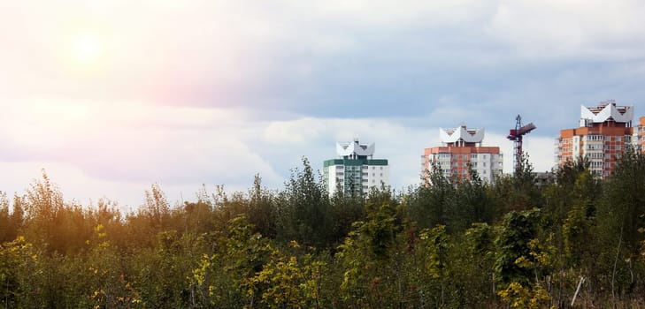 Nestled within a vibrant green area, a collection of modern buildings with distinctive rooftops emerges against a backdrop of soft, cloudy skies. The golden hues of dusk bathe the scene in a warm glow, contrasting the natural foliage that surrounds the urban structures.
