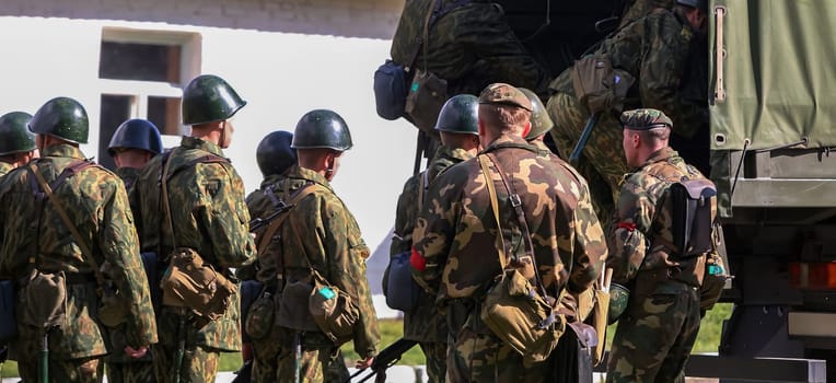 A group of soldiers in camouflage uniforms and helmets gathers near a military truck, preparing for transport during a training exercise at a base.