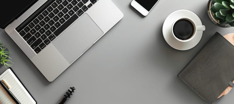 An overhead view of a modern, minimalist workspace featuring a laptop, smartphone, coffee cup, stylish journal, pen, and small potted plant on a clean, gray desk. ideal for illustrating productivity and organization in a sleek, professional setting.