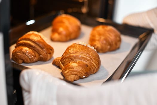 female baker taking tray with fresh croissants from oven in modern kitchen, close-up