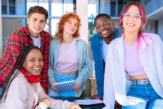 A group of five diverse students studying and collaborating at a library table working on a shared project, with books and devices on the table.
