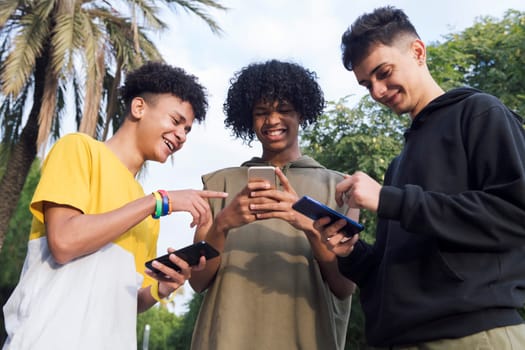 low angle of teen diverse friends having fun surfing Internet on cellphones smiling happy in park, concept of youth lifestyle and technology of communication