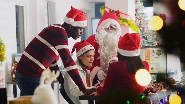Team leader dressed as Santa during Christmas season delegates project tasks to workers. Director in xmas decorated workspace discussing with employees during winter holiday season