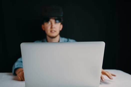 A young man with a VR headset is focused on using a laptop in a dimly lit room, showcasing his hightech engagement
