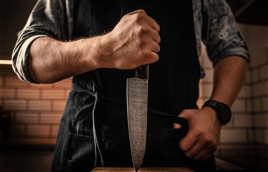 Chief man in apron holding knife vertically at kitchen stubbing wooden board. Restaurant proffesional with culinary tool closeup