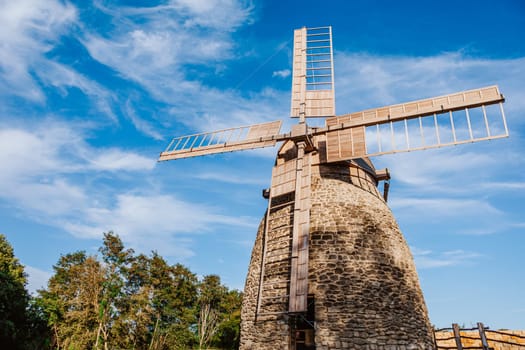 Old stone windmill stands against a clear blue sky with gentle clouds. The windmill wooden blades and stone structure showcase traditional craftsmanship and the historical significance of rural architecture.
