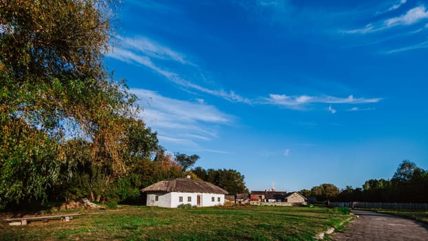 Peaceful rural scene featuring a traditional thatched-roof house and a wide open road under a vast blue sky. The whitewashed house stands out against the greenery of trees and fields, creating a picturesque countryside setting.