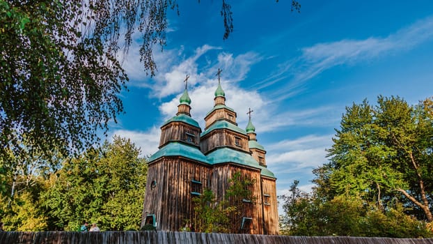 Traditional wooden church with green domes rises behind a rustic wooden fence, set against a beautiful blue sky with wispy clouds.