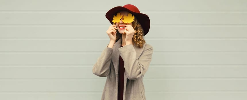 Autumn portrait of beautiful happy smiling young woman covering her eyes with yellow maple leaves, stylish joyful girl wearing hat on gray background