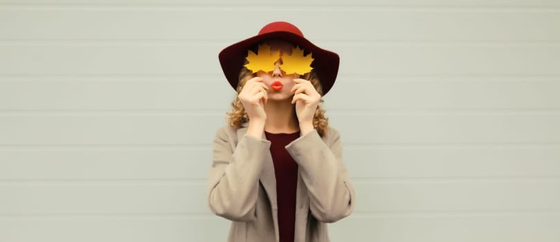 Autumn portrait of beautiful happy young woman covering her eyes with yellow maple leaves, stylish joyful girl wearing hat on gray background