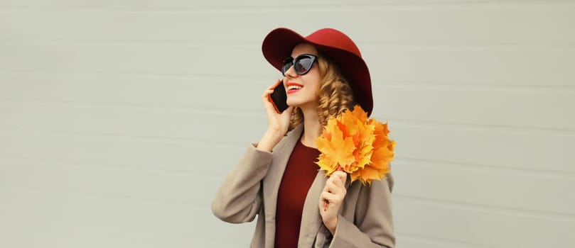Autumn portrait of happy smiling young woman calling on phone, stylish girl talking on smartphone holding yellow leaves in elegant hat looking away on gray background