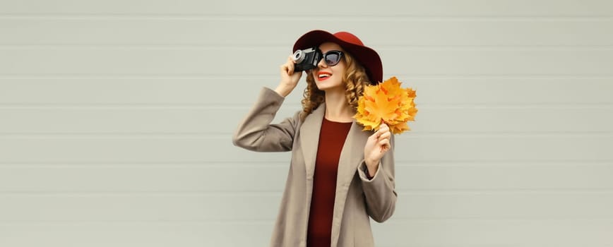 Autumn portrait of beautiful happy young woman photographer taking picture with camera, stylish girl holding yellow maple leaves looking away wearing hat on gray background