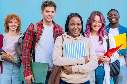 Group of smiling diverse students on university campus with notebooks in hands standing on blue background looking at camera