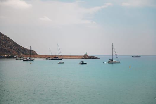 A peaceful harbor scene featuring several boats anchored in calm, turquoise waters under a serene blue sky, with a rocky coastline in the background.