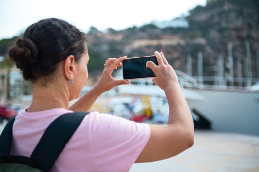 A woman takes a photo of a picturesque marina using her smartphone, highlighting travel and leisure activities.