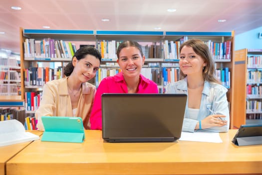 Three girl students with laptops and notes sit in the library to review the exam looking at camera