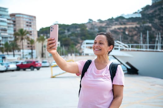 Happy woman enjoying a sunny day by the marina, taking a selfie with her smartphone. She wears a casual outfit and backpack, capturing memories of her travel experience.