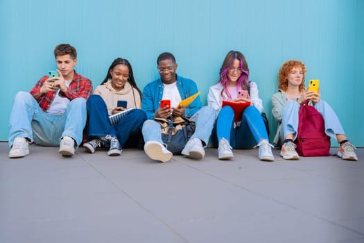 A diverse group of five smiling young students sit together on the floor, smiling and interacting on their smartphones against a blue background.