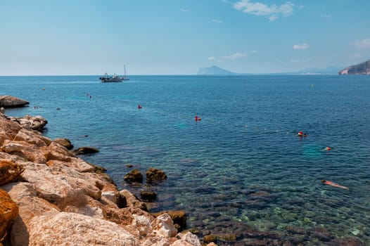 Beautiful coastal scene featuring clear blue waters, swimmers enjoying the sea, rocky shore, and a boat on the horizon under a clear sky.
