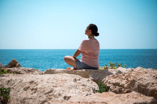 A woman sits cross-legged on rocky terrain, meditating peacefully with an expansive view of the blue ocean, embracing the tranquility and serenity of the natural surroundings.