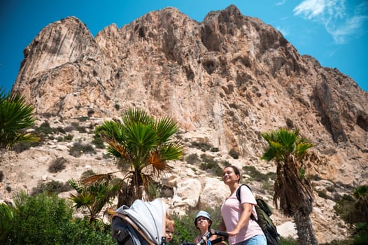A family enjoys a hiking adventure beneath a towering rocky mountain. A parent pushing a stroller stands amidst lush greenery, palm trees, and clear blue skies.