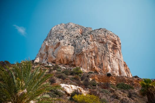A stunning view of a towering rocky cliff under a clear blue sky, surrounded by lush green vegetation, representing nature's beauty and serenity.