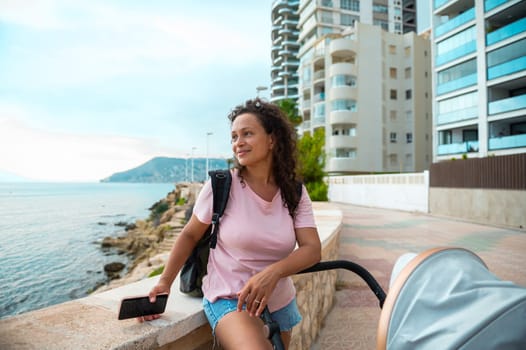 Smiling woman sitting by the sea, enjoying a sunny day with her baby in a stroller. Urban backdrop enhances the leisurely vibe.