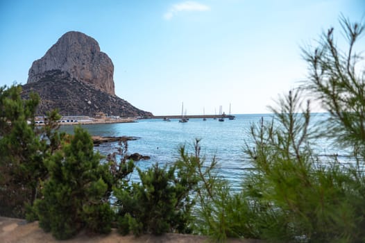 Scenic view of Penon de Ifach in Calpe, Spain, with serene waters and sailboats. Lush greenery frames the tranquil Mediterranean coastline, creating a perfect summer escape.