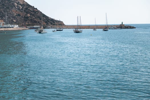A peaceful coastal scene with several sailboats anchored in a calm marina, with a backdrop of a rocky mountain and open sea under a clear sky.
