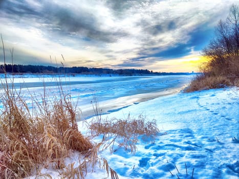 The frozen river reflects the colorful sunset while grasses emerge from the snow, creating a peaceful winter landscape.