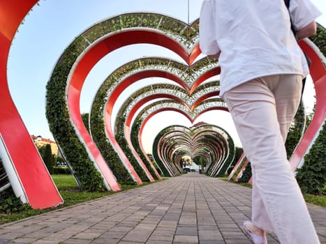 A person strolls beneath heart-shaped arches covered in greenery, enjoying an evening in a lively park setting.