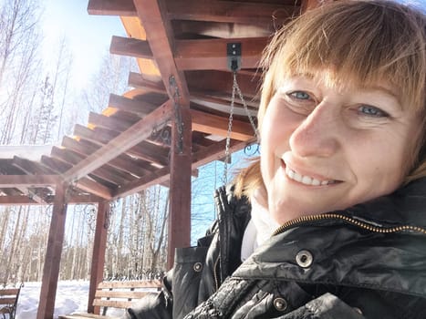 A cheerful middle aged woman in a winter coat taking selfie on the swing on nature outdoors in sunny day with blue sky