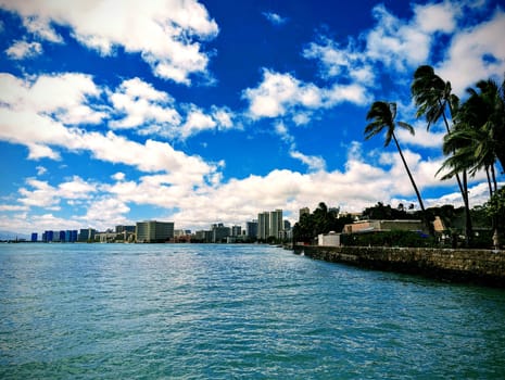 A vibrant view of Waikiki featuring palm trees, calm water, and a skyline, all set against a backdrop of fluffy clouds and deep blue sky.