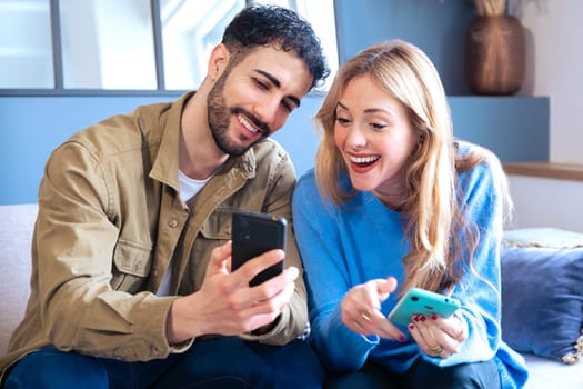 A young smiling multiracial couple sits on the couch at home using a cell phone browsing a wireless internet application on a device. Technology concept.
