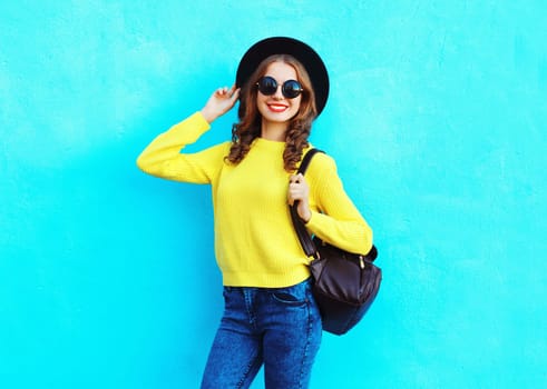 Stylish happy smiling woman in black hat, yellow knitted sweater and backpack on blue background