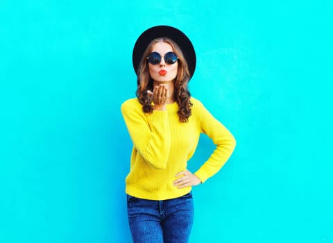 Fashion beautiful woman blowing kiss in black hat, yellow sweater on colorful blue background