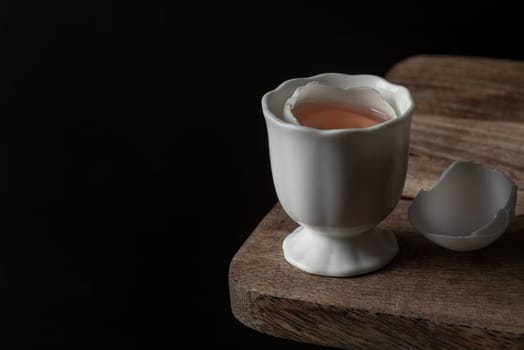 Yolk and White egg in A cracked raw white duck egg shell in White porcelain egg cup on the edge of a wooden cutting board with dark background. use it as your Wallpaper, Poster and Space for text, Selective focus.
