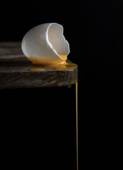 A cracked raw white duck egg shell and dripping yolk on the edge of a wooden cutting board and  black background. Conceptual image of a broken white eggshell with dripping egg yolk on dark background, use it as your Wallpaper, Poster and Space for text, Selective focus.