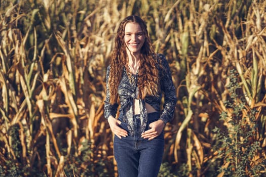 In a picturesque cornfield, a joyful young woman stands confidently in a striking pose, her bright smile radiating warmth under the golden glow of the sunlight, surrounded by tall, lush cornstalks
