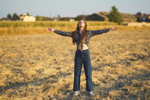A young woman stands in a beautifully sunlit field, her arms outstretched wide as a symbol of her freedom and pure joy. The golden hues of nature gently envelop her in a serene, idyllic outdoor scene