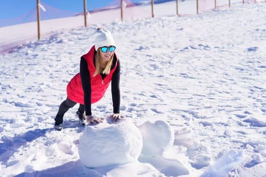 Full body of smiling young female blonde in beanie cap polarized sunglasses, looking away while leaning on hands and doing push ups on rock against wooden pillars fence in daylight